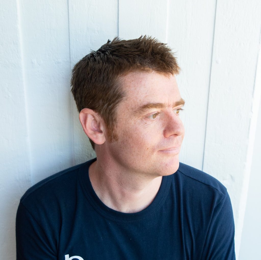 Picture of Matt Chittock in a navy t-shirt, against a white backdrop, smiling, looking away.