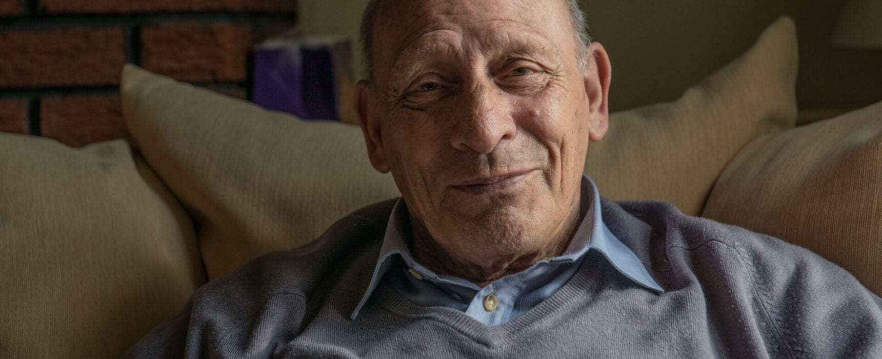 An older man smiling, sitting on an armchair at home.